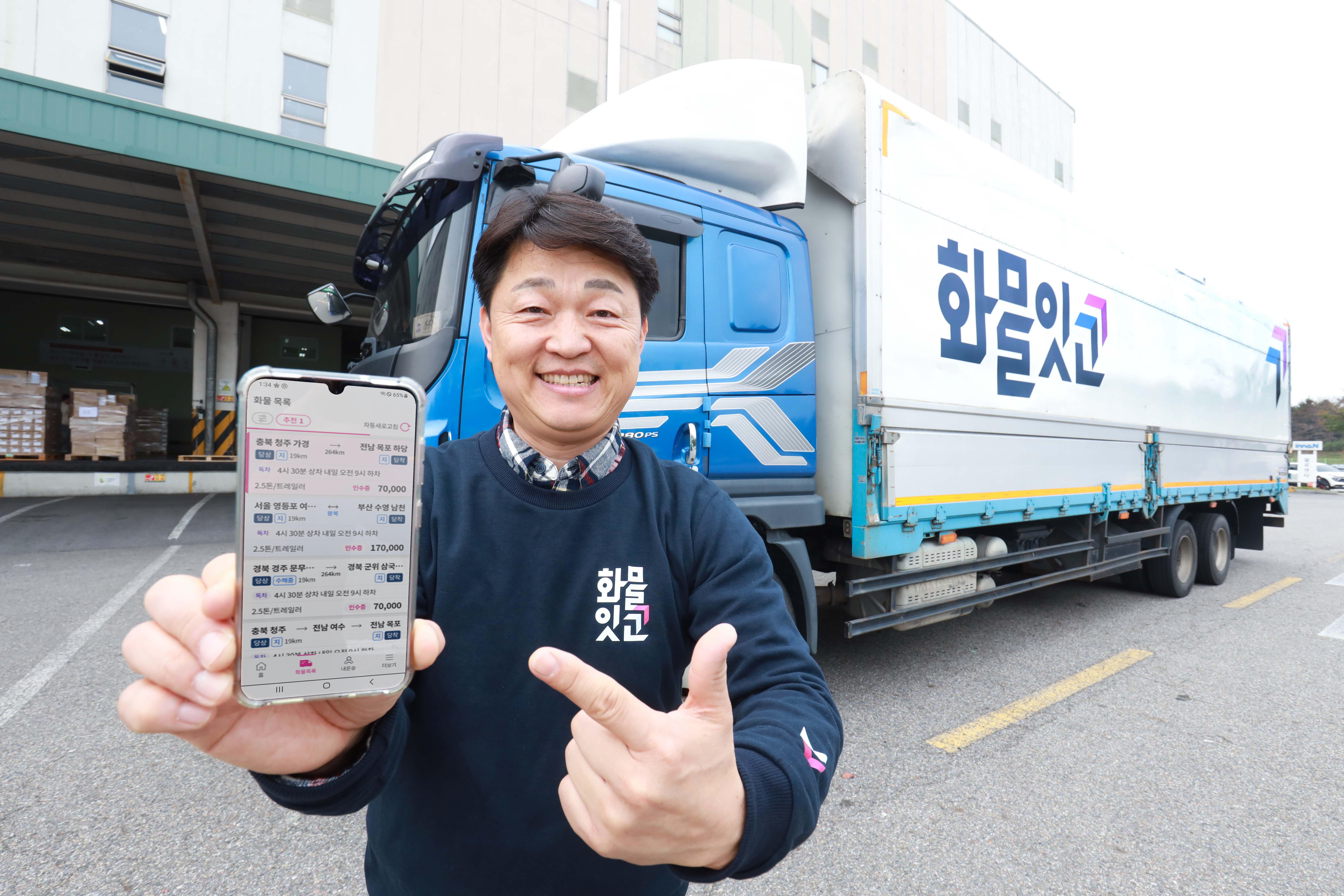 파란 대형 화물차 앞에서 '화물잇고' 로고가 있는 티셔츠를 입은 남성이 스마트폰을 들고 웃고 있는 이미지